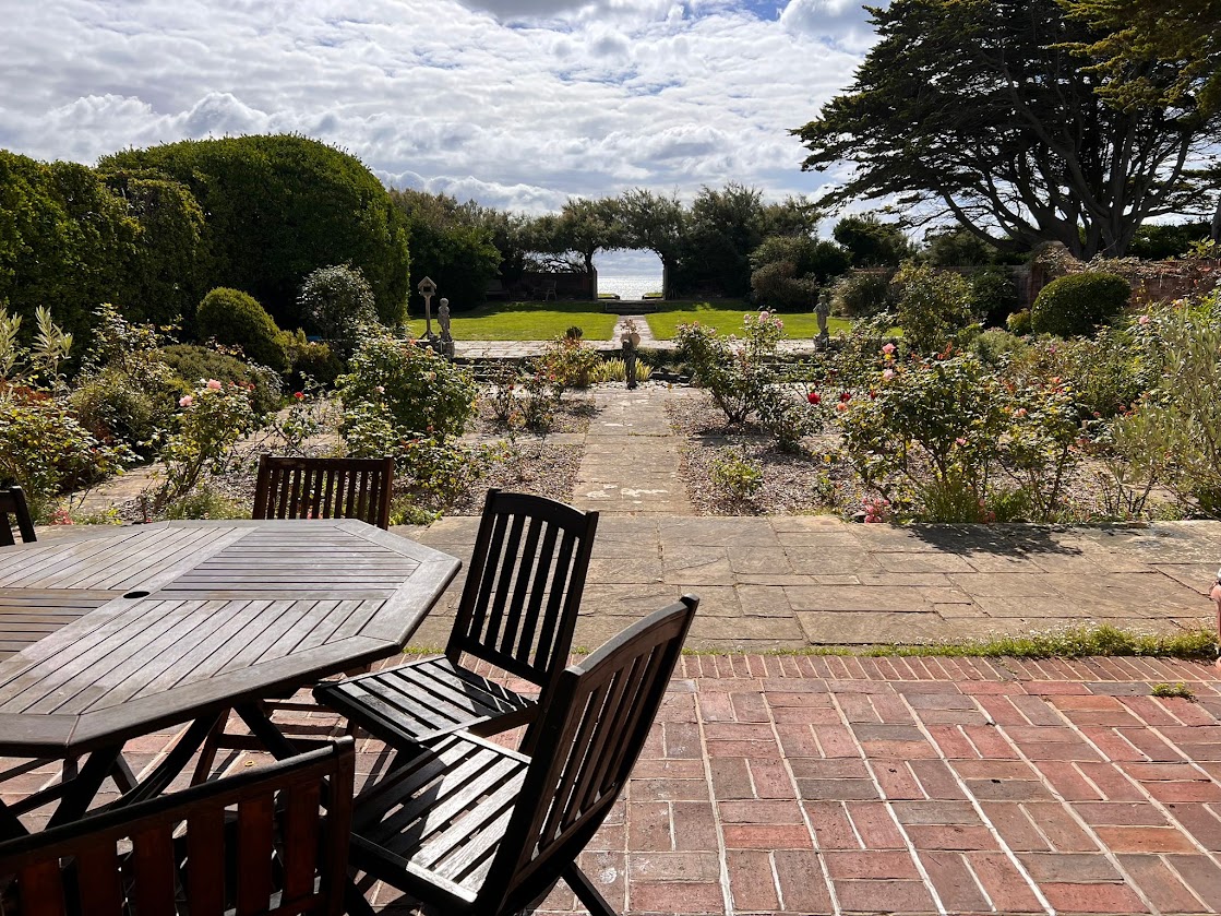 The view from the covered outdoor seating area at Blue Waters, with a wooden table and chairs on the brick terrace looking over the rose garden towards the sea