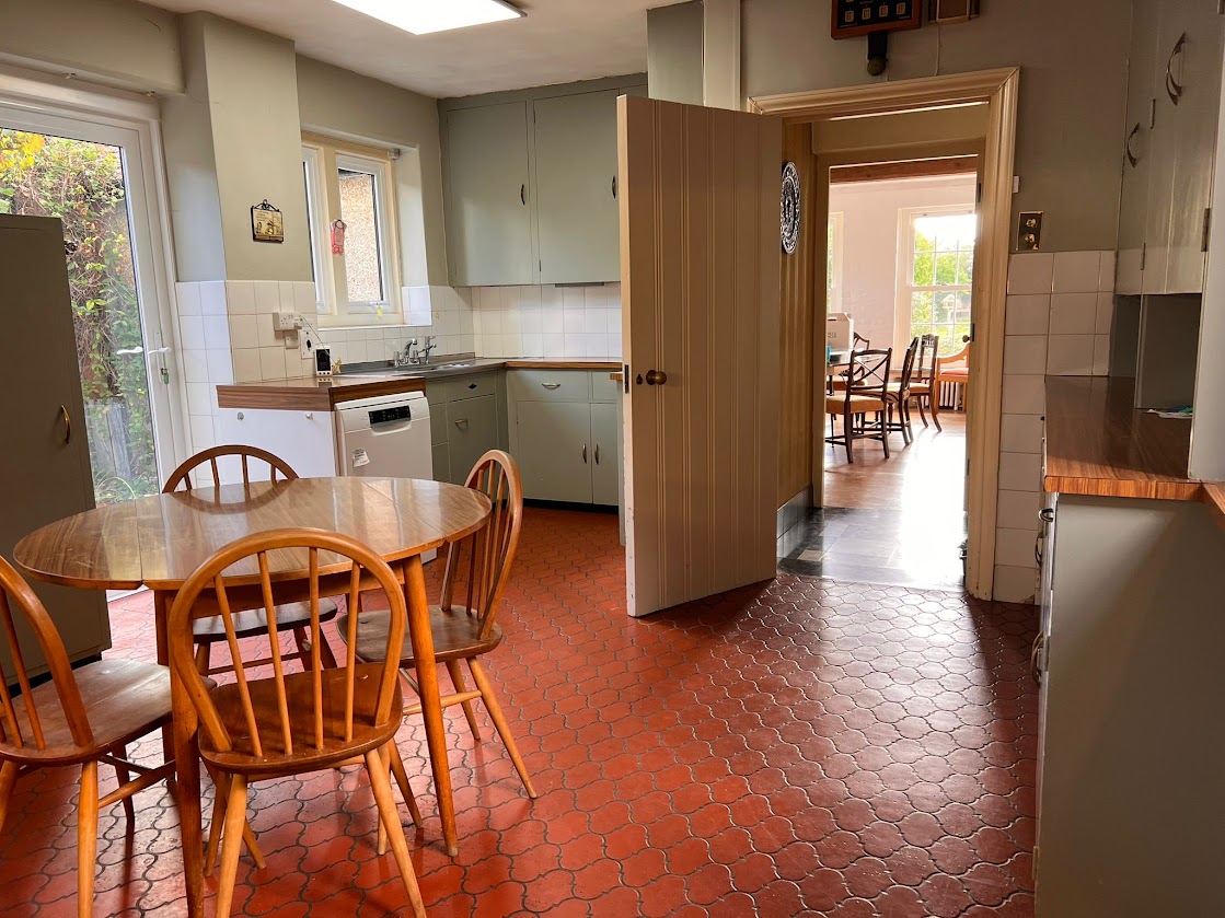 A second view of the kitchen at Blue Waters with the round table, sage-green cabinets and terracotta floor looking through to the dining room
