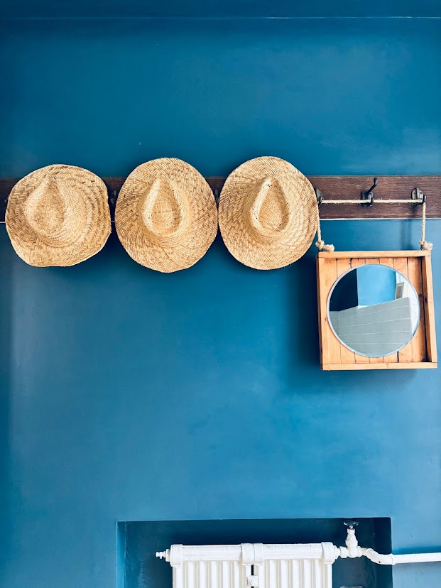Straw hats hanging on a wooden rack against a deep blue wall in the hallway at Blue Waters